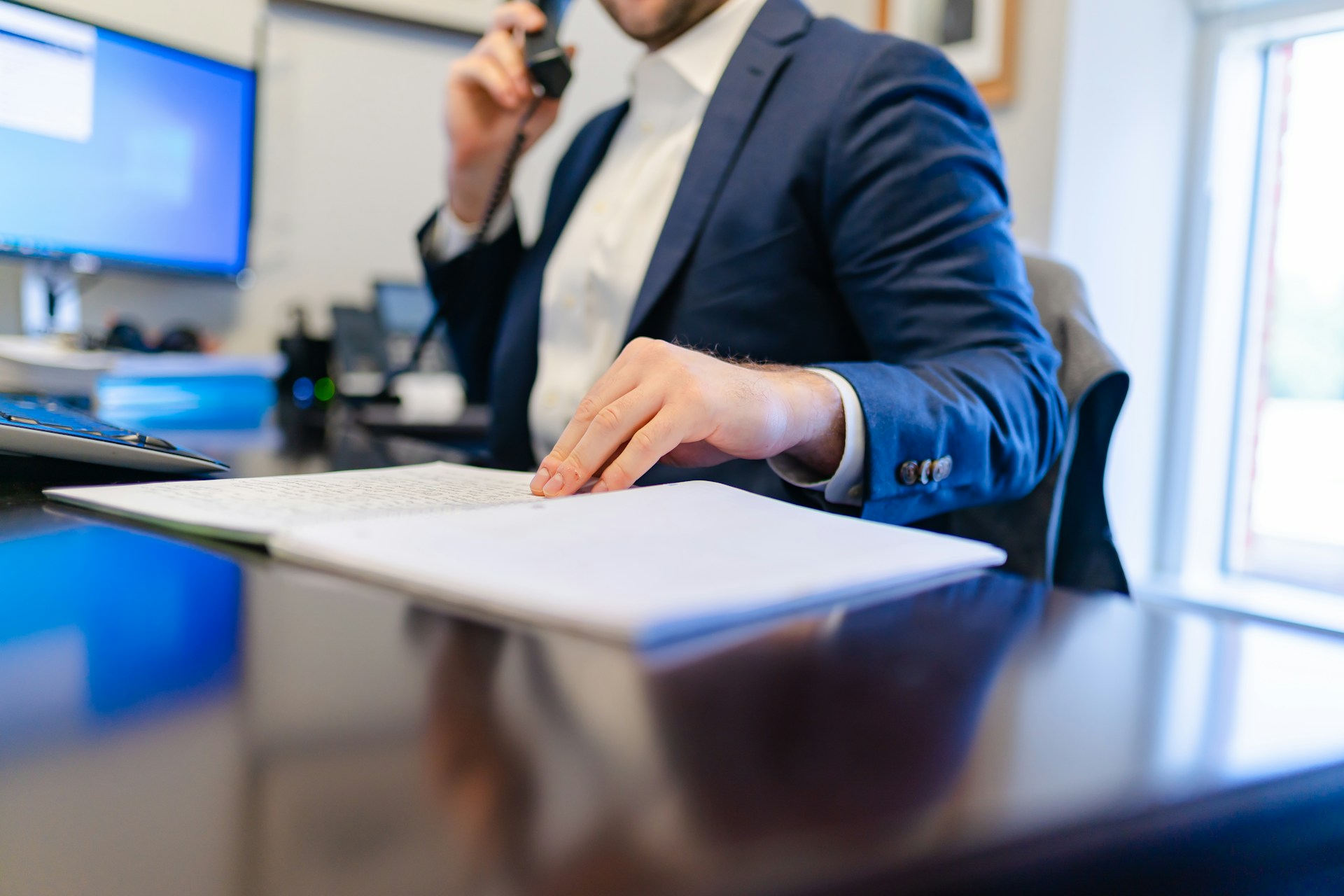 An advisor in a blue suit resting a finger on an open printed file at a polished desk while holding a phone to his ear
