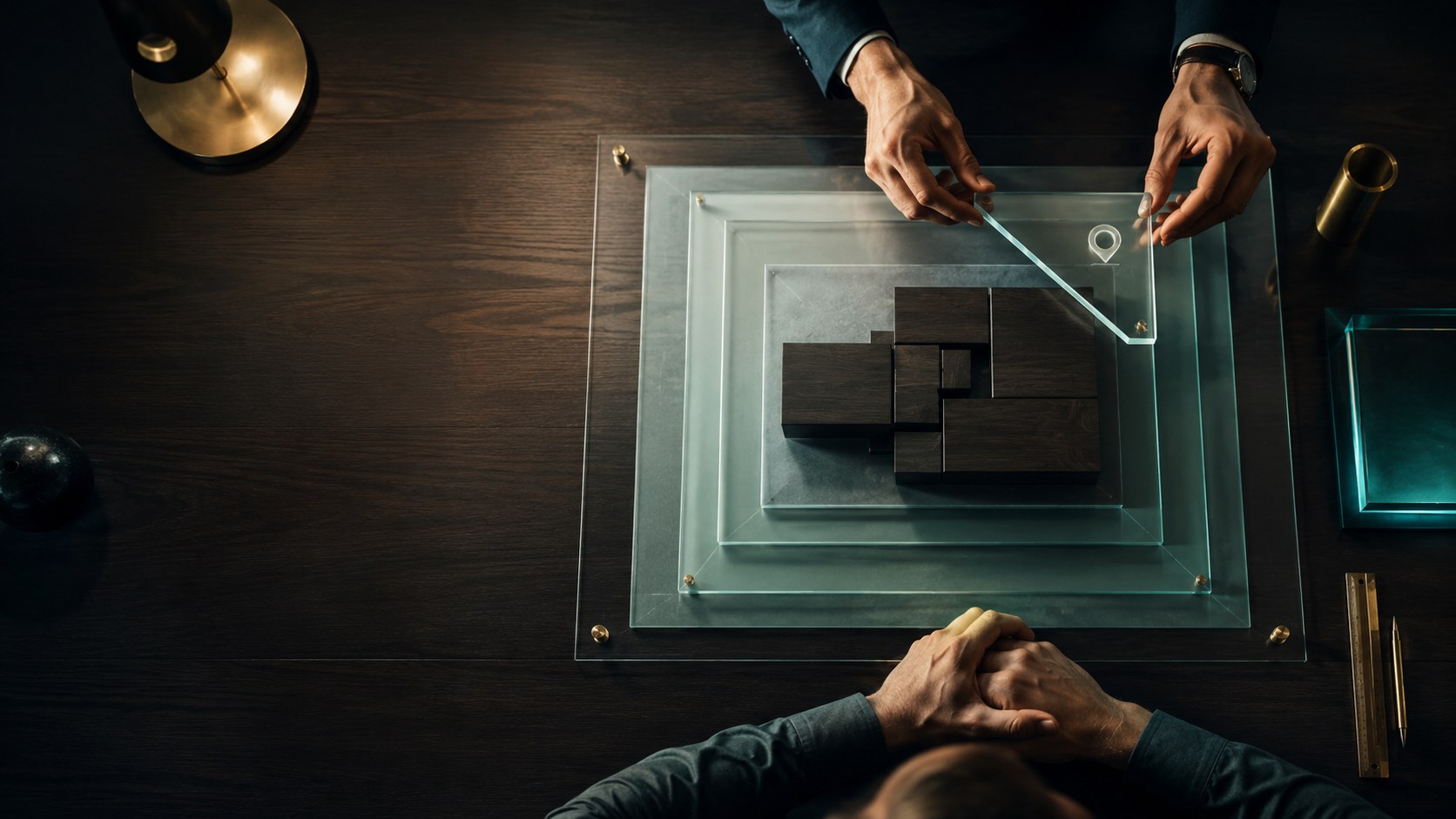 Hands lifting an etched glass plate from a stack of frosted plates carrying dark massing blocks on a walnut desk